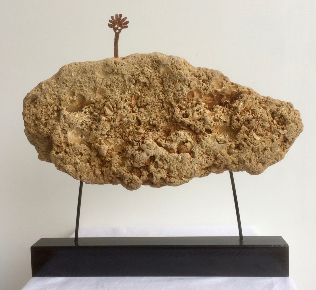 A sculpture of Rock Spirit - a copper stick figure with head, eyes and mouth and large spicy hair but no limbs - the Spirit stands on top of a piece of yellowish Coraline Crag, a fossiliferous limestone from Suffolk.